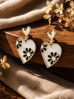Gold-toned heart dangle earrings with black and white enamel floral detail, laid flat